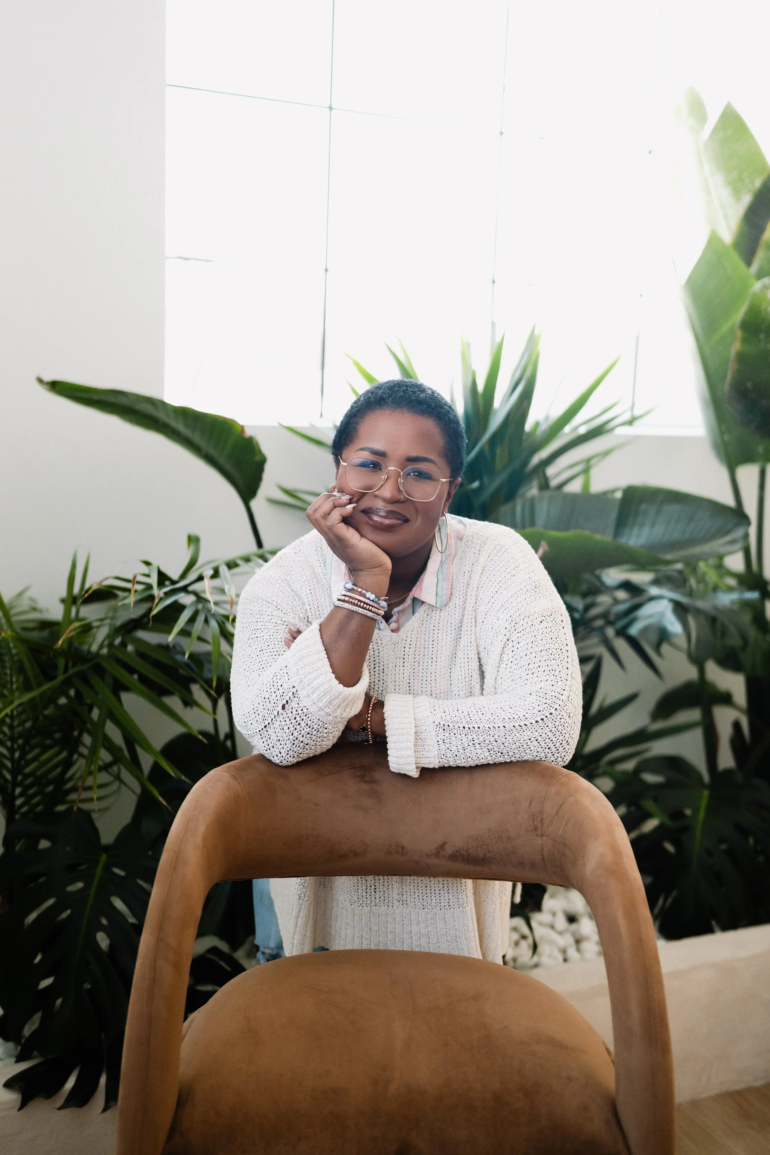A woman with short curly hair, glasses, and dark lipstick sits at a table with her chin resting on her hand, smiling. She is surrounded by large green plants and sits in front of a window with white blinds that let in natural light.