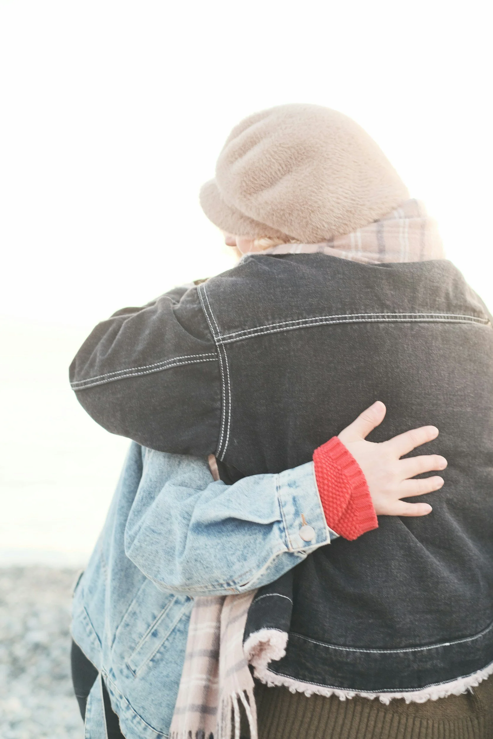 Two people hugging each other, one person wearing a tan knit beanie, a gray and beige plaid scarf, and a dark denim jacket, and the other wearing a light blue denim jacket with a red thumb cuff, and a beige scarf with fringes. The background is bright and blurred.