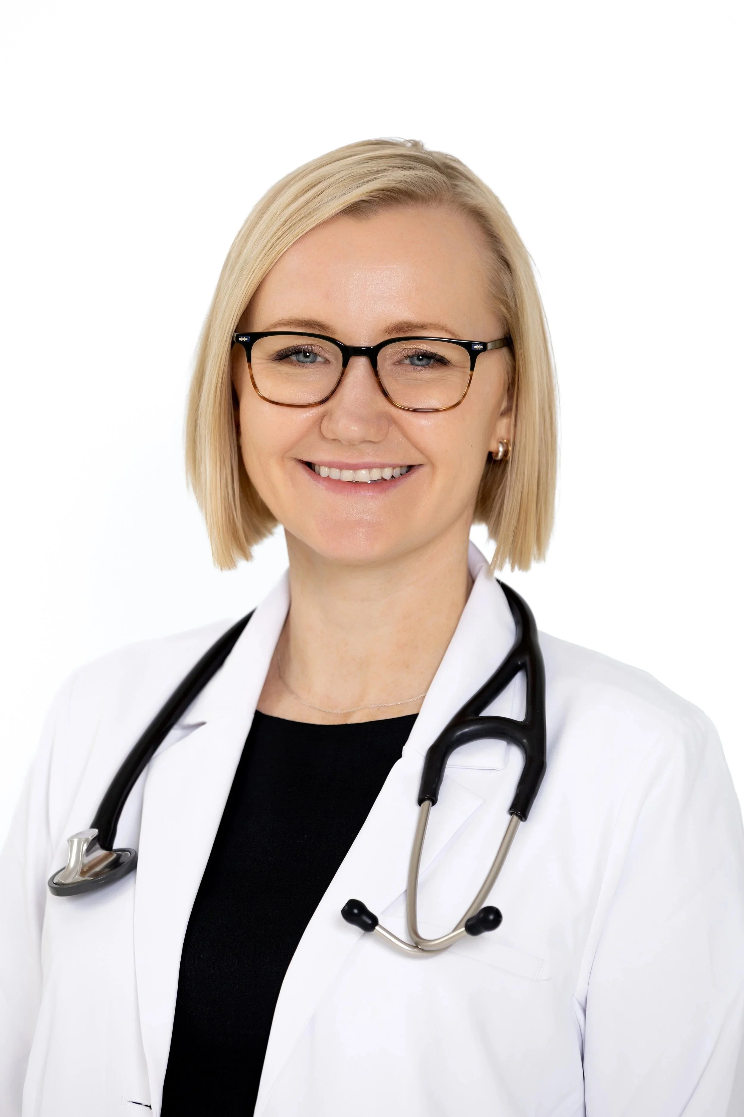 A smiling female doctor with blonde hair, glasses, and a stethoscope around her neck, wearing a white coat and black shirt, against a white background.