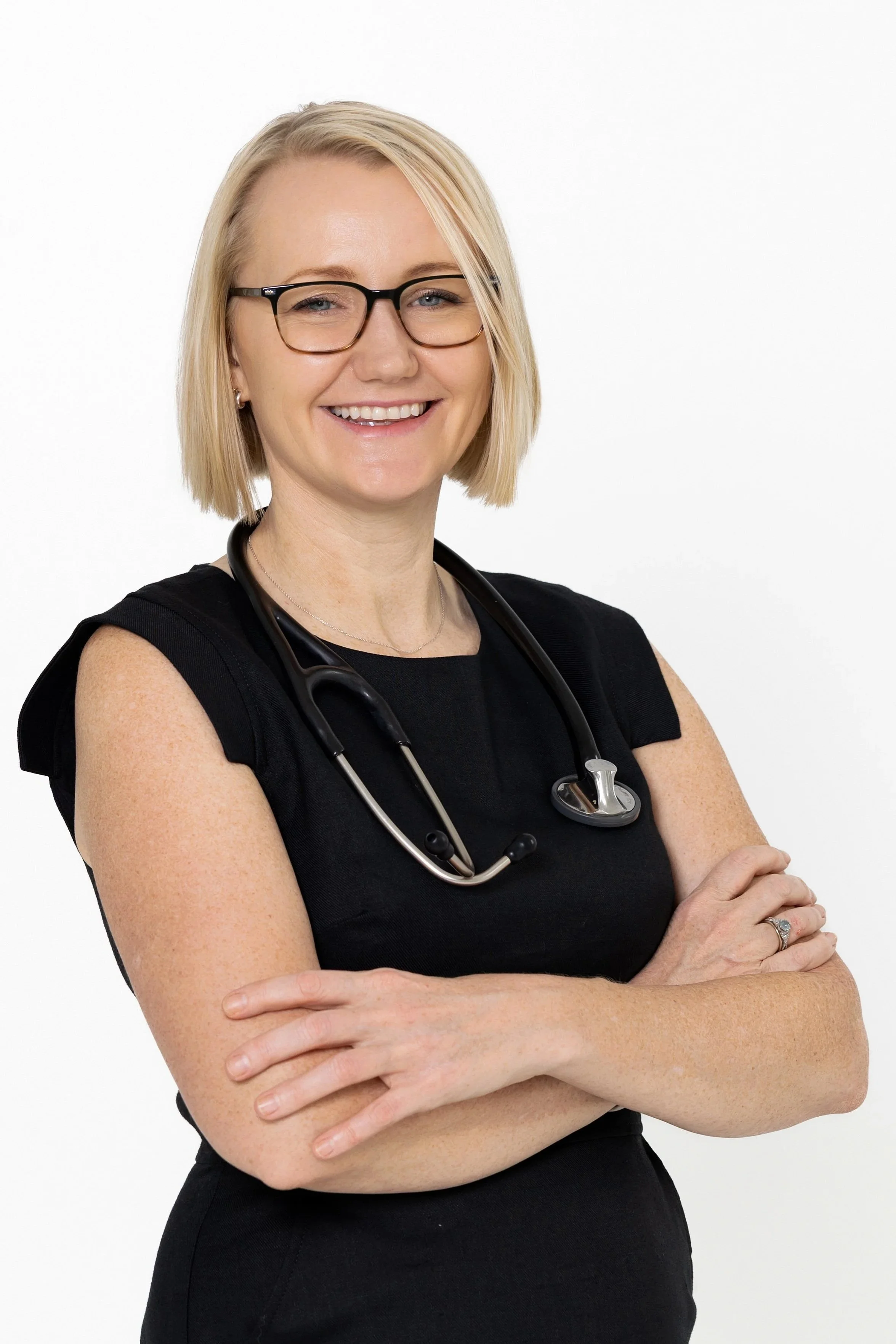 A smiling female healthcare professional with blond hair, wearing glasses, a black sleeveless top, and a stethoscope around her neck, standing with her arms crossed against a plain white background.