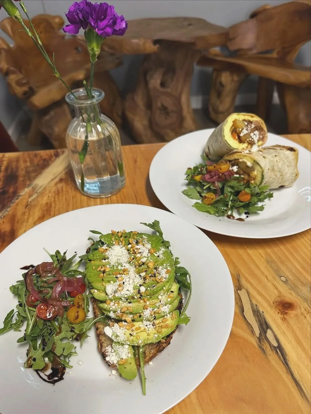 A wooden table with two plates of food: one with an avocado toast topped with cheese, seeds, and herbs, and a side of salad; the other with a wrap cut in half, filled with vegetables and possibly meat, accompanied by a small salad. There is a small glass vase with purple flowers in the background.