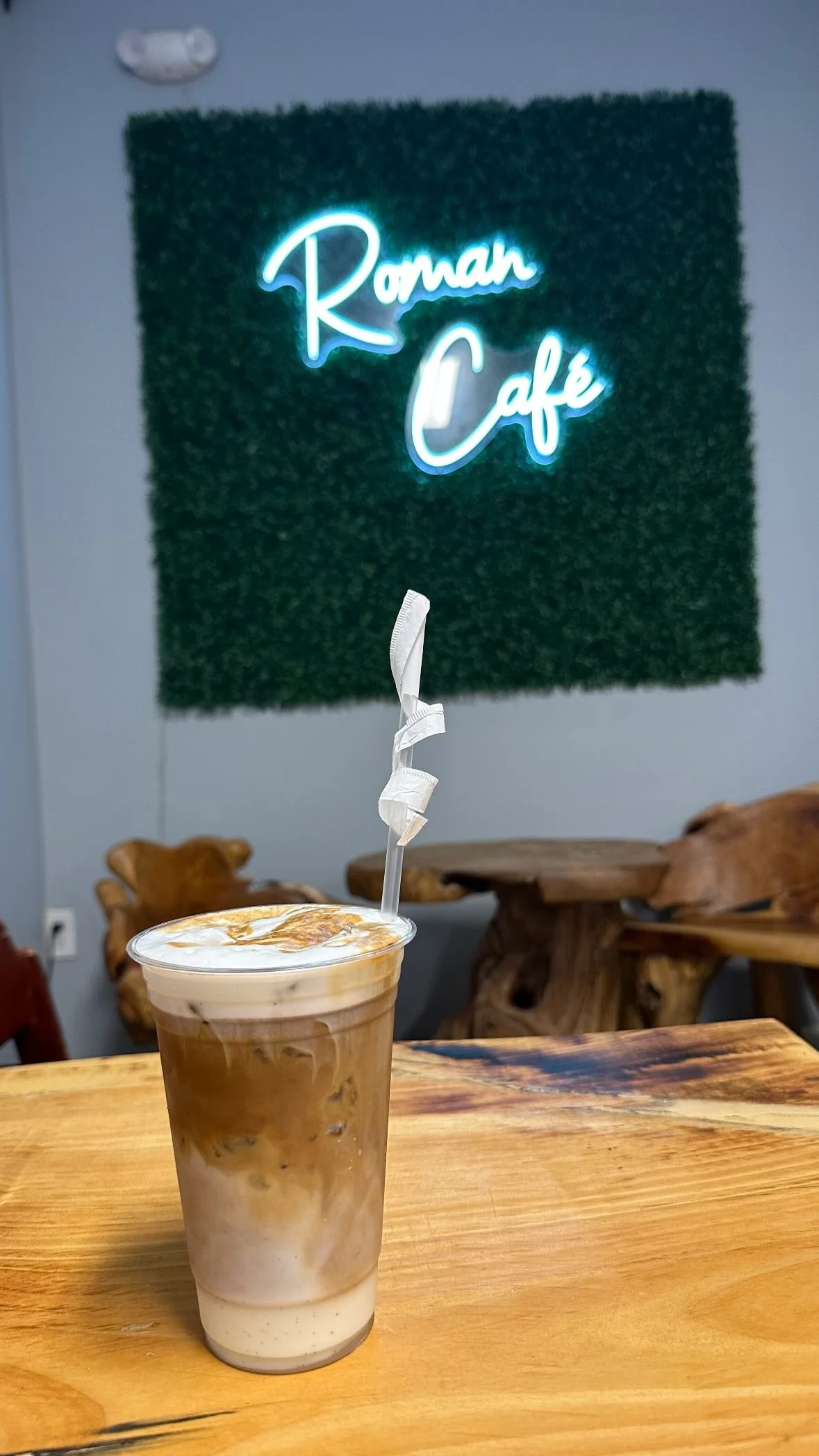 A plastic cup filled with iced coffee topped with caramel drizzle, placed on a wooden table in a cafe with a neon sign that says 'Roman Cafe' on the wall behind.