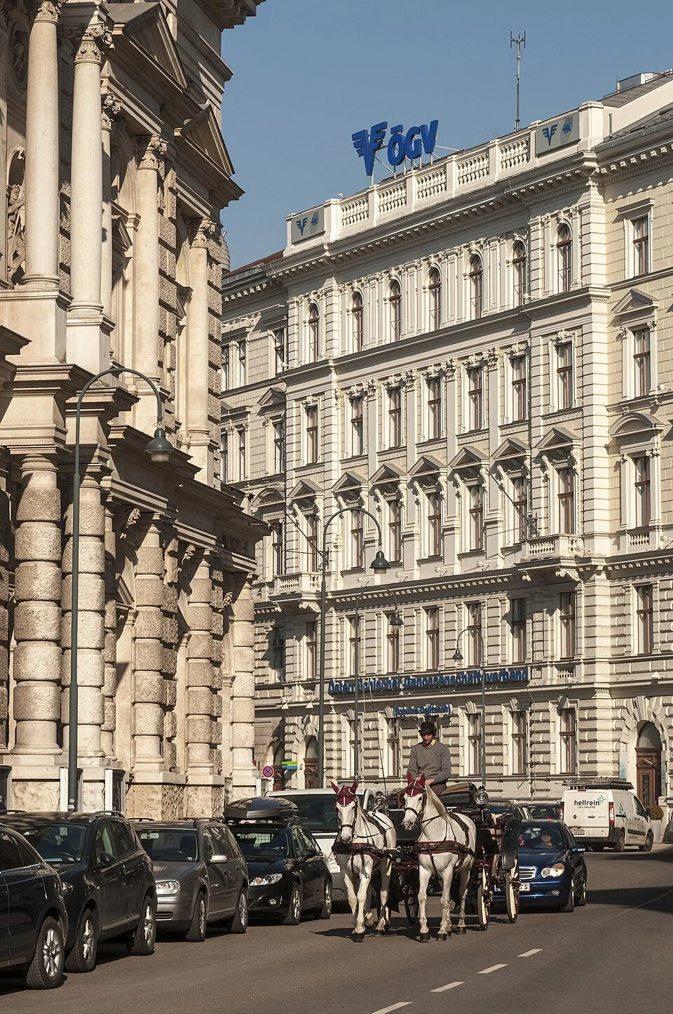 A city street scene with historic buildings, parked cars, a horse-drawn carriage, and a person driving the carriage. The buildings have ornate architectural details and signs on the rooftops.