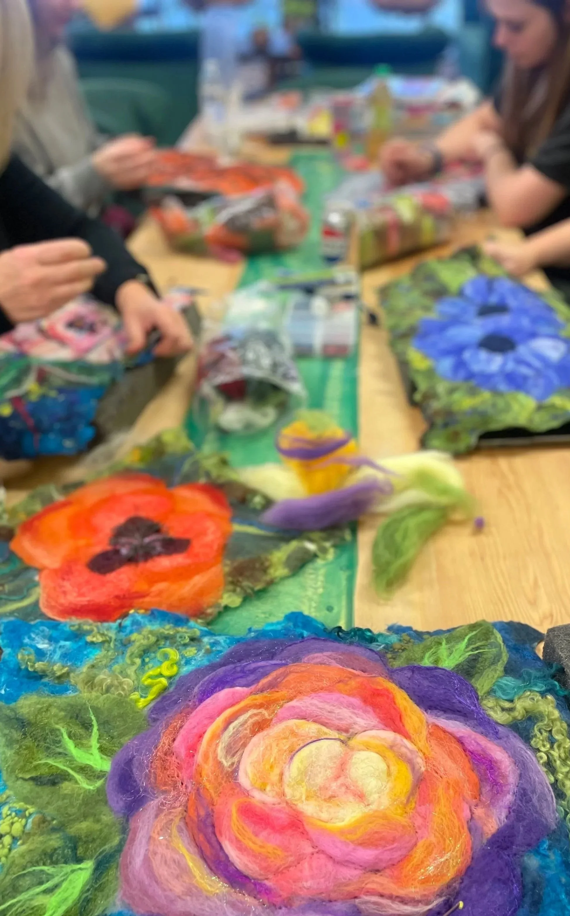 Colorful felted wool art pieces resembling roses in vibrant pink, purple, orange, and yellow hues are displayed on a table in a craft room with people crafting in the background.
