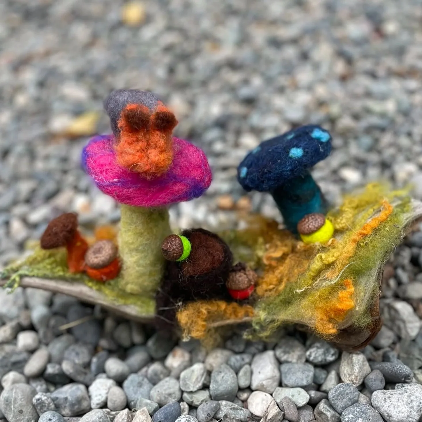 Colorful felted mushrooms with stems and caps in pink, blue, orange, and black, surrounded by felted leaves and small felted berries, placed on a bed of small gray stones.