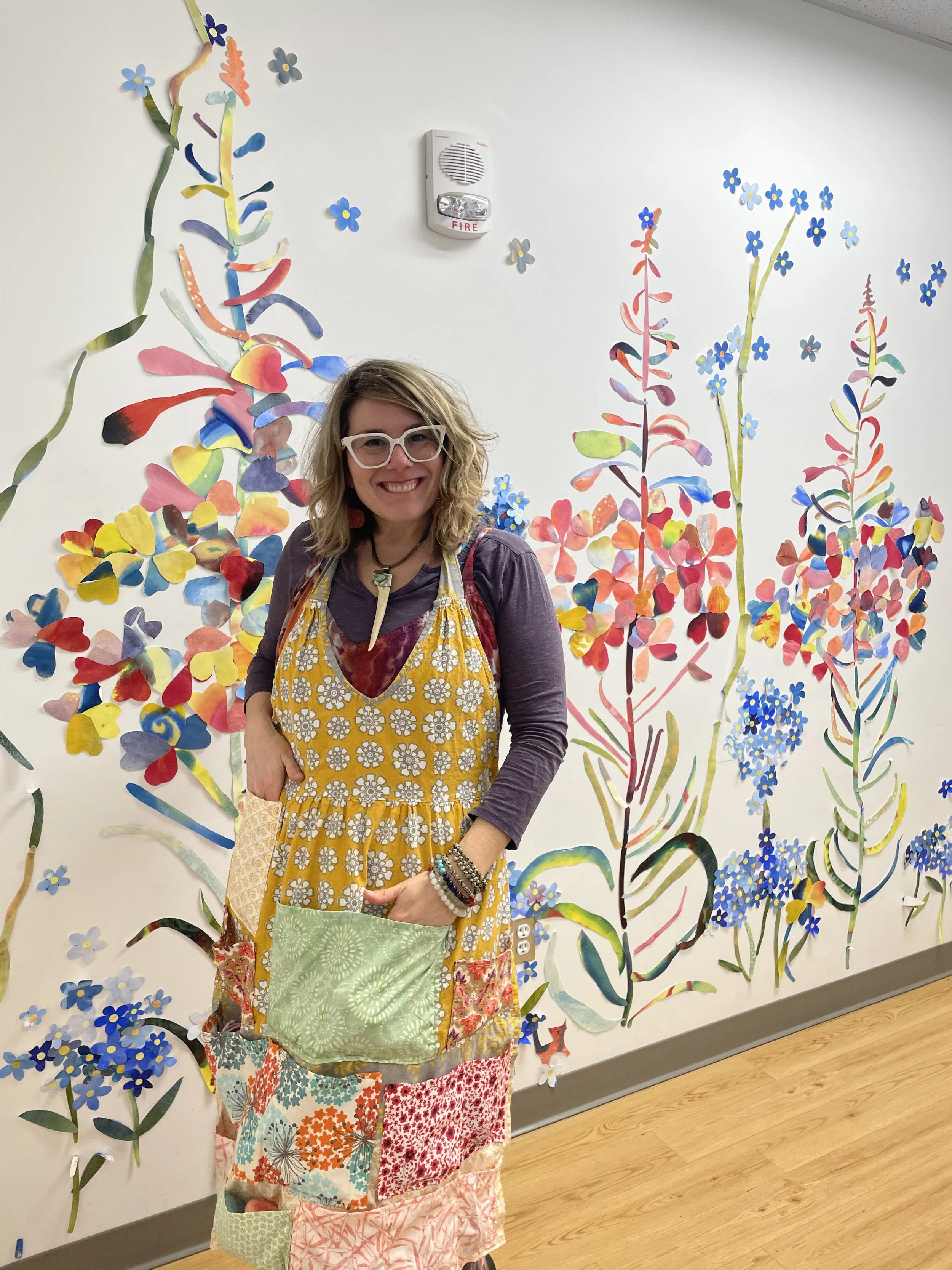 A woman with curly hair wearing glasses, a purple top, a yellow apron with flower patterns, and multiple bracelets, standing in front of a colorful art wall featuring large floral and foliage designs made of paper or fabric, in an indoor setting with wooden flooring.