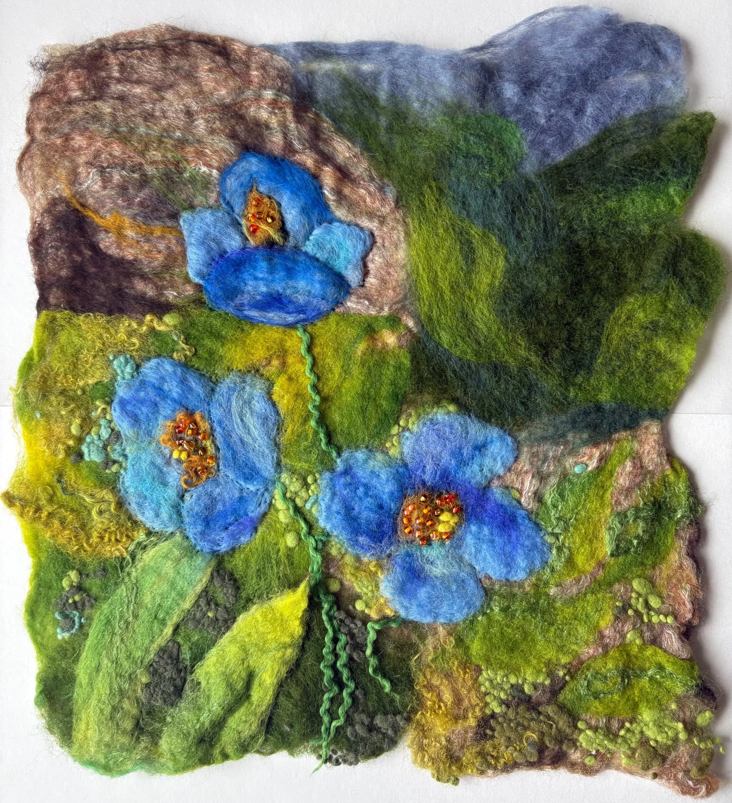 Felted art depicting blue flowers with yellow and red centers on a green and brown background.