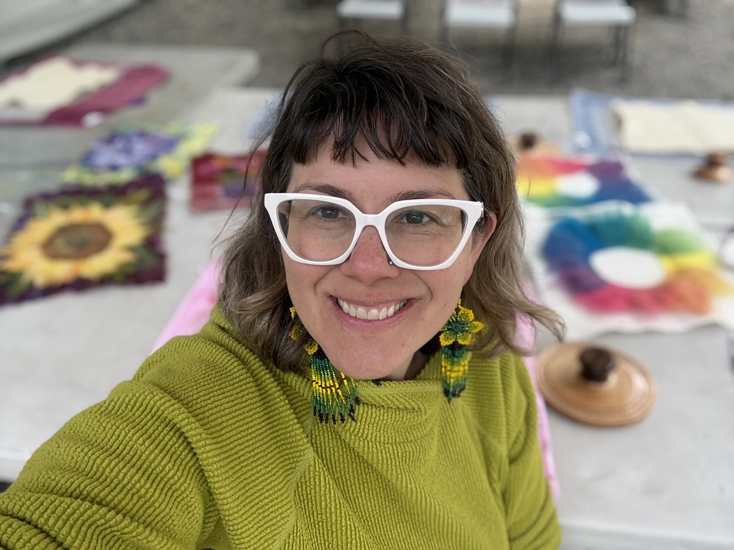 A smiling woman with brown hair, wearing white glasses, a green sweater, and colorful beaded earrings, taking a selfie at an arts and crafts fair with rainbow-colored crafts in the background.