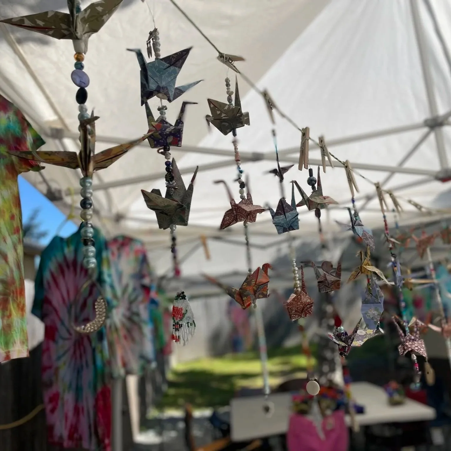 Colorful origami cranes hanging on strings under a canopy at an outdoor market or craft fair.