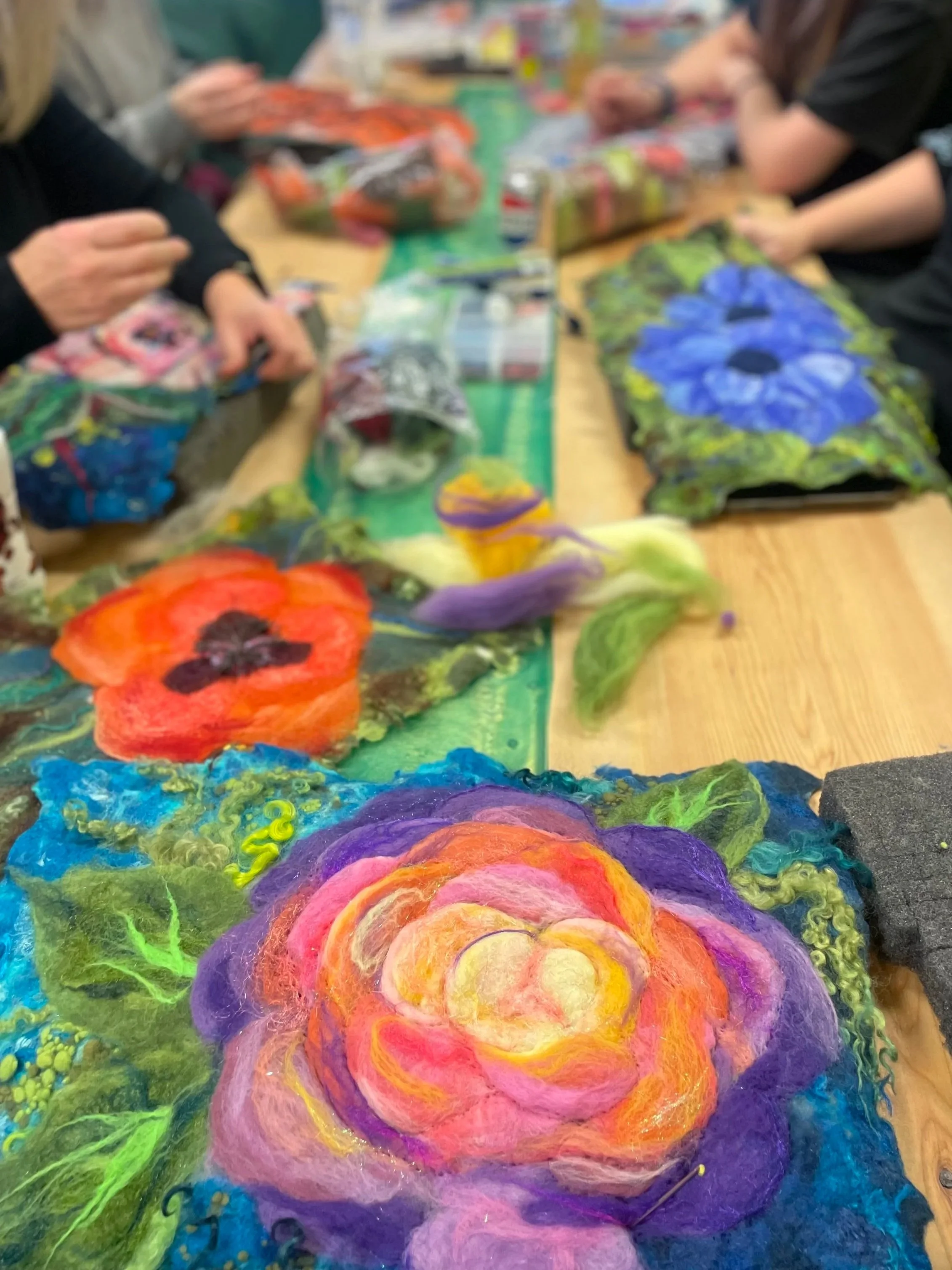 Close-up of vibrant felted wool art depicting a multicolored flower with layers of pink, purple, orange, and yellow petals, surrounded by green leaves and a vivid multicolored background, on a table at an art or craft workshop.