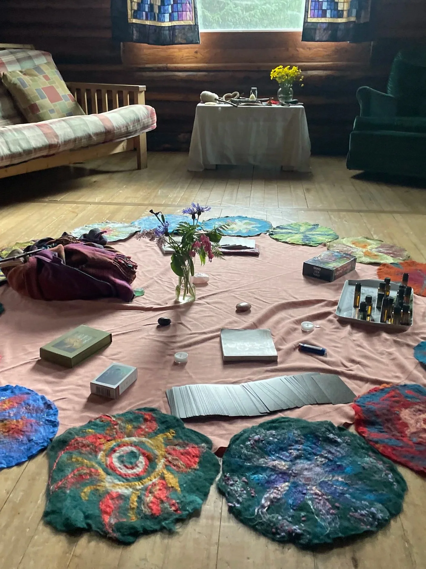 A room with various colorful, circular fabric or yarn art pieces laid out on a pink sheet on the wooden floor. There is a vase with flowers, books, and other items on the sheet, and furniture such as a plaid sofa, a green armchair, and a small table with a white cloth and yellow flowers in a vase. The room has wooden walls, a window with curtains, and a natural, cozy atmosphere.