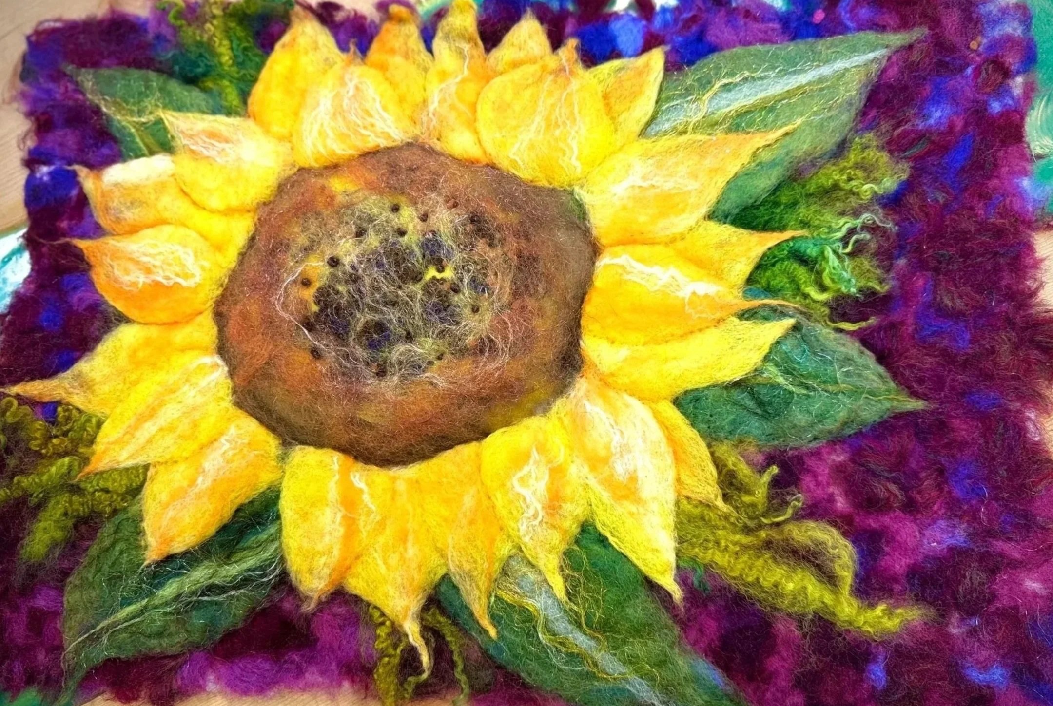 A vibrant, colorful felted artwork of a sunflower with bright yellow petals and a brown center, surrounded by multicolored purple, green, and blue felted leaves and background.