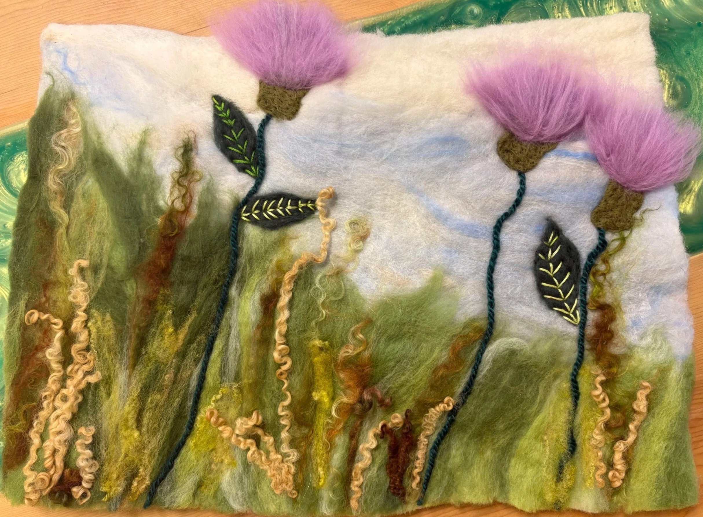 Close-up of felted wool artwork depicting a nature scene with trees and leaves, with pink and brown felted mushrooms or flowers at the top.