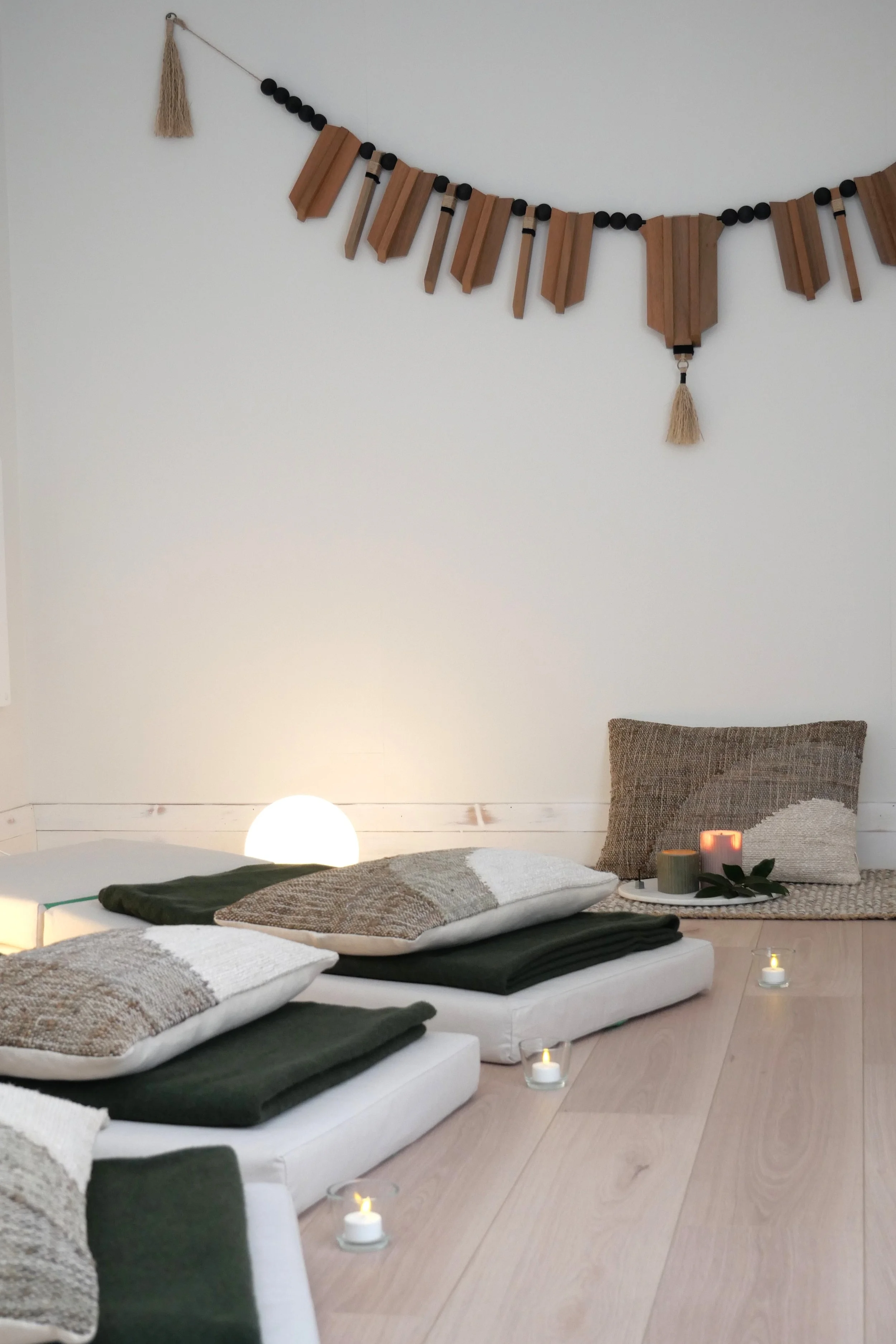 A cozy indoor space set up for relaxation or meditation with cushions, low tables, candles, and decorative items, against a white wall with a hanging wall art piece.