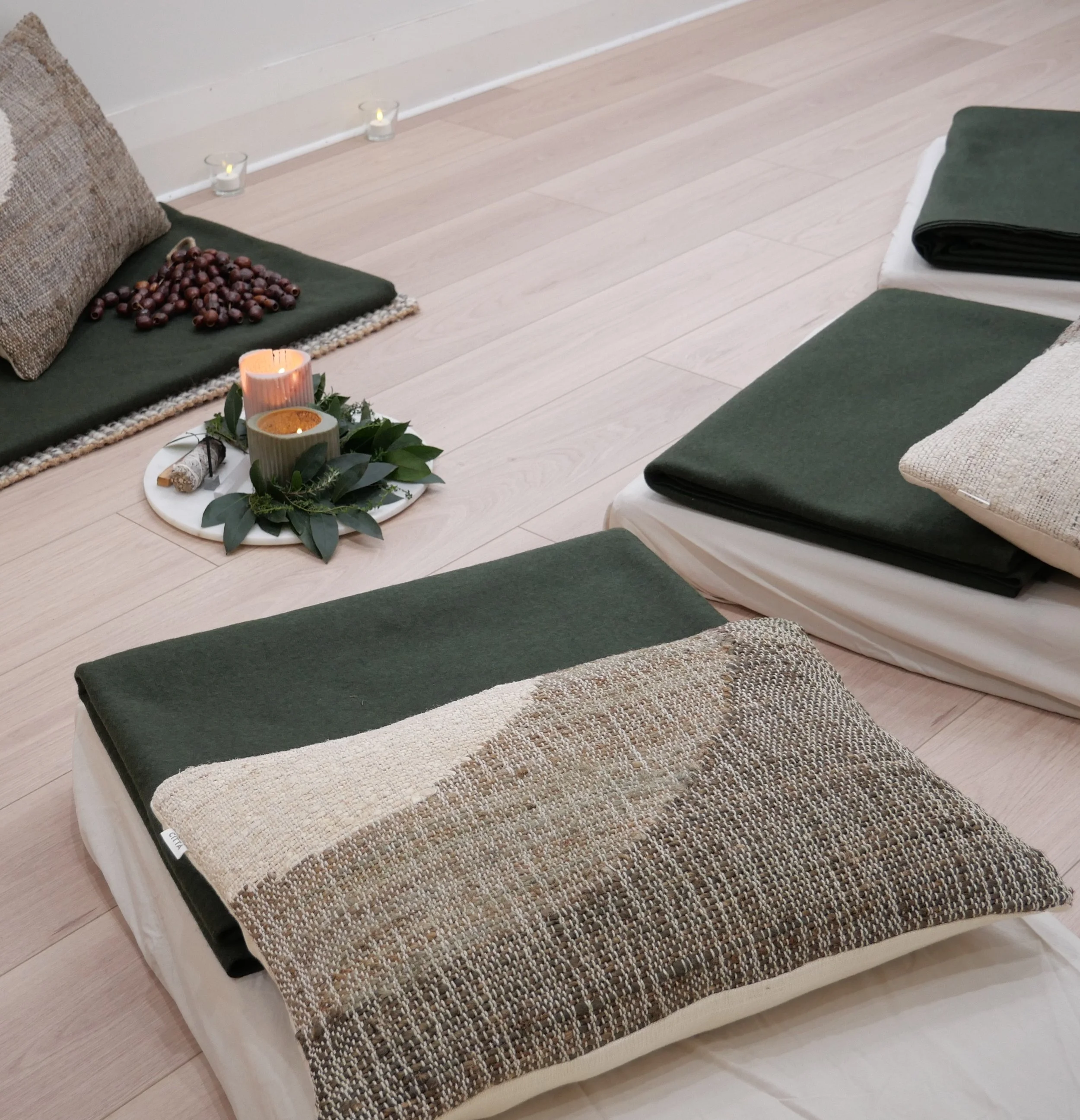 A cozy indoor setting with a cushioned seating area, decorative candles, and a tray of greenery and pinecones. The seating features a woven textured pillow and a dark green cushion, set against a light-colored wooden floor.