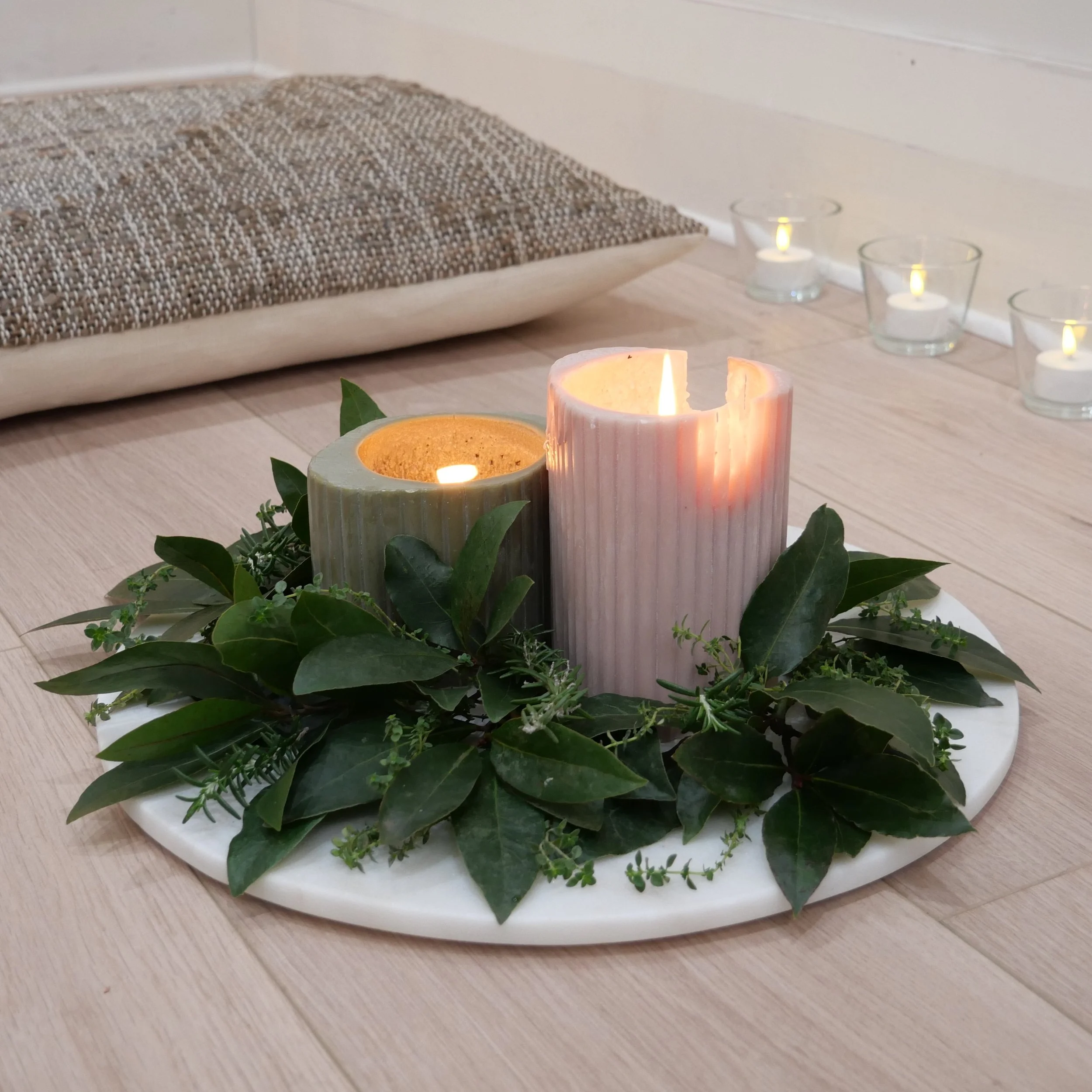 Sandstone candle holders with lit candles surrounded by green leaves and small plants, on a light wood surface.