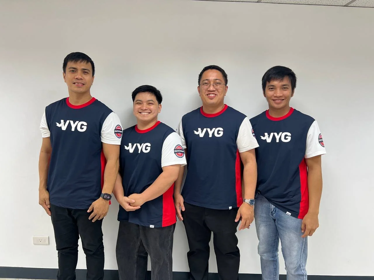 Group of four men standing against a plain wall, all wearing matching blue, white, and red sports jerseys with logos.