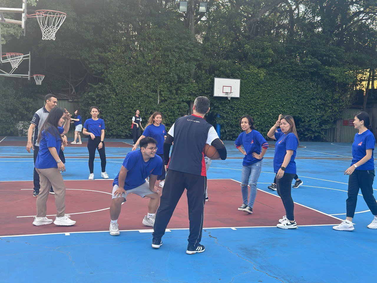 A group of people on an outdoor basketball court, engaging in a basketball activity with a coach holding a basketball, surrounded by trees.