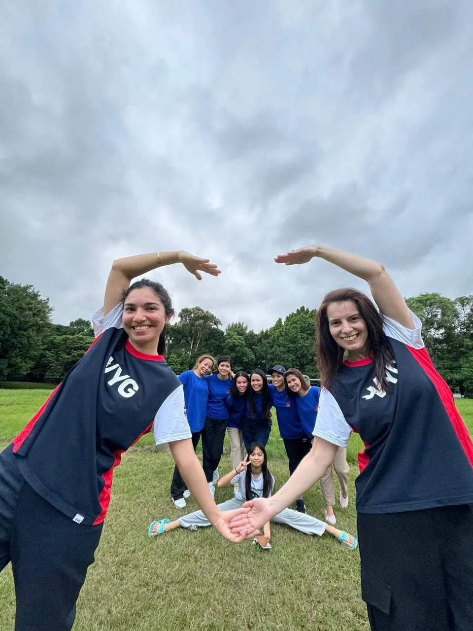 Group of women and girls outdoors, forming a circle, with two women in the front holding hands above their heads. The group is on a grassy field with trees and cloudy sky in the background.