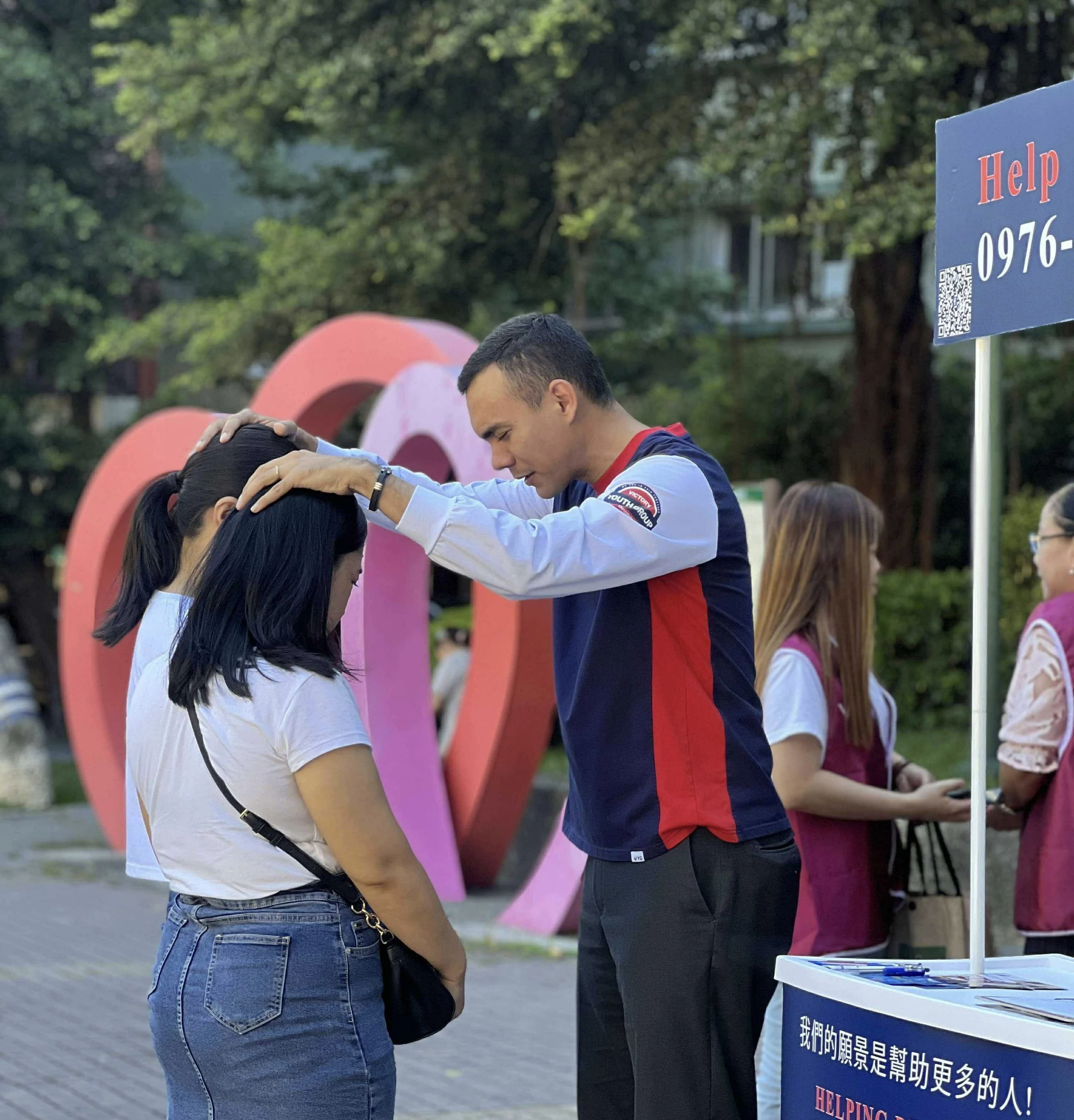 A man in a blue and red shirt is performing a religious act of laying hands on a woman in a white shirt with black hair, during an outdoor event with a crowd and a help signboard with Chinese text in the background.