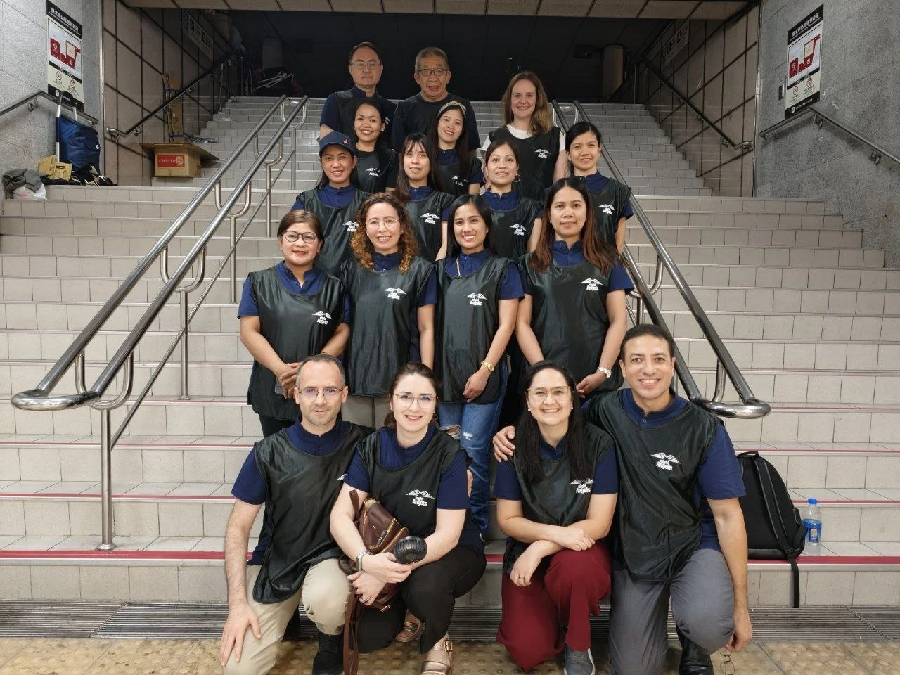 A group of people posing on stairs at an indoor location, most are wearing matching black vests over blue shirts. They are smiling and standing in front of a grey wall with signs and metal railings.
