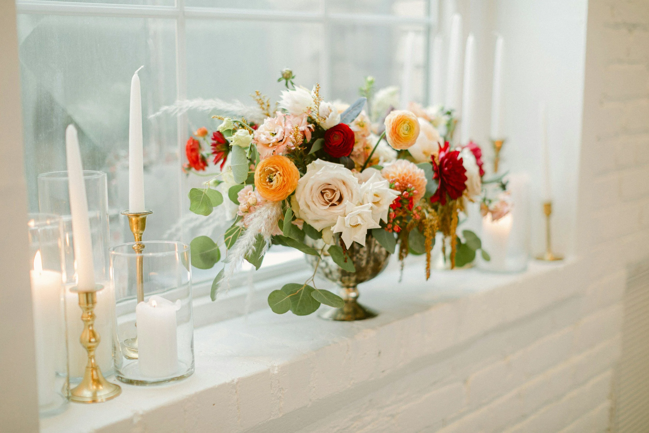 A floral arrangement with various flowers, including roses and ranunculus, placed on a windowsill surrounded by white candles in glass holders, in a well-lit room with white walls.