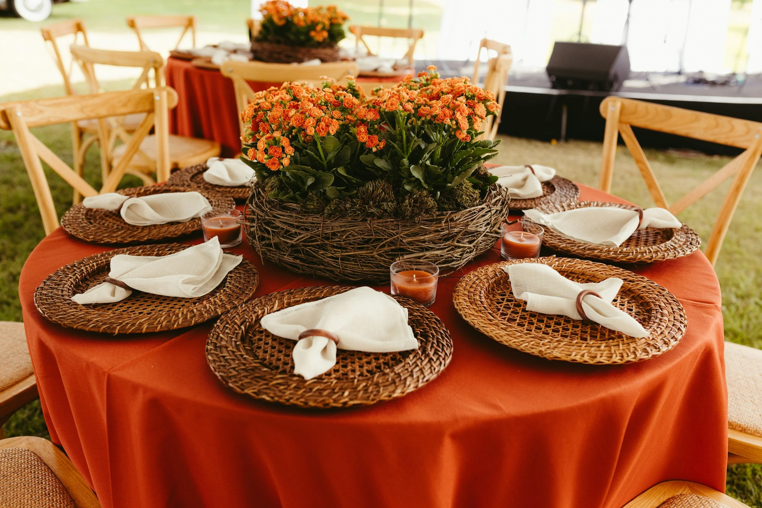 Round table with an orange tablecloth, set for a meal with woven placemats, white napkins, and small candles. A large basket with orange flowers decorates the center of the table. Wooden chairs surround the table, and there is a stage with audio equipment in the background.