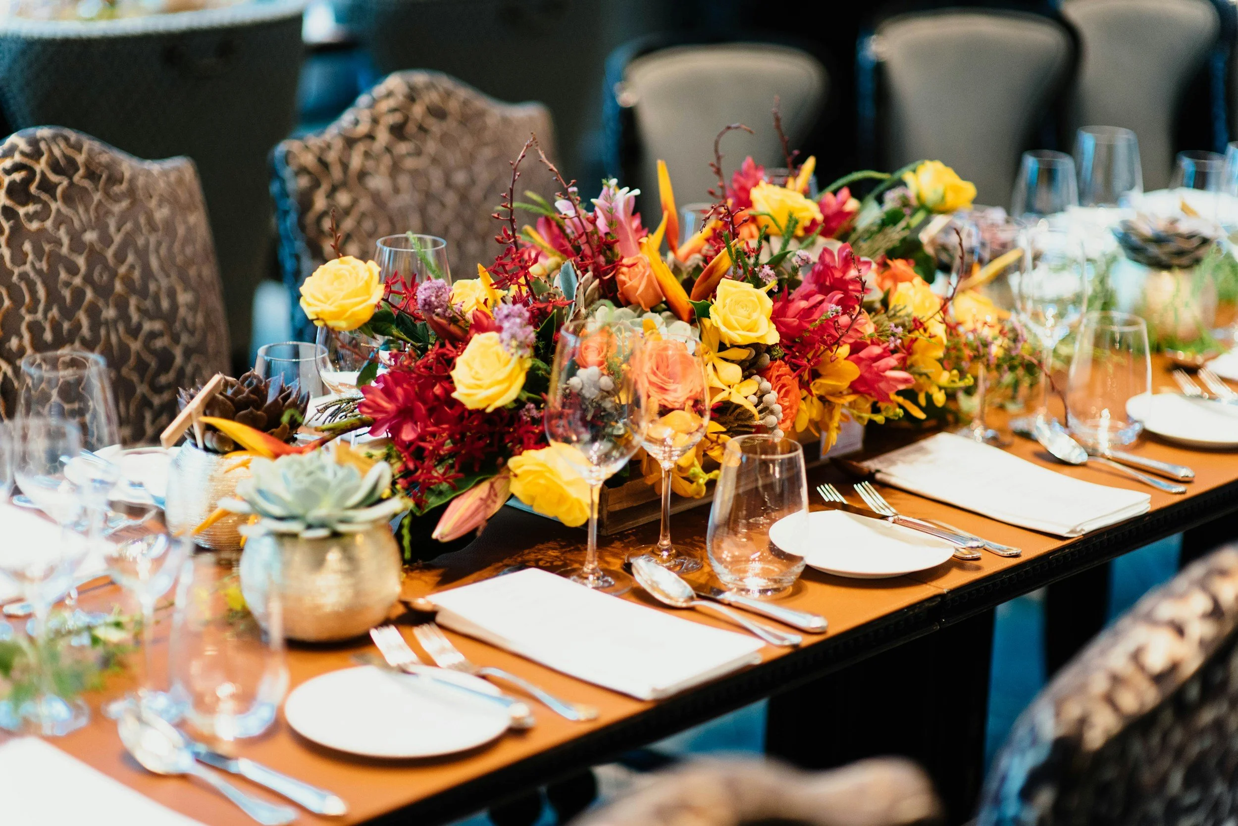 A long dining table decorated with a colorful centerpiece of various flowers, surrounded by glassware, plates, and cutlery, set for a formal event.