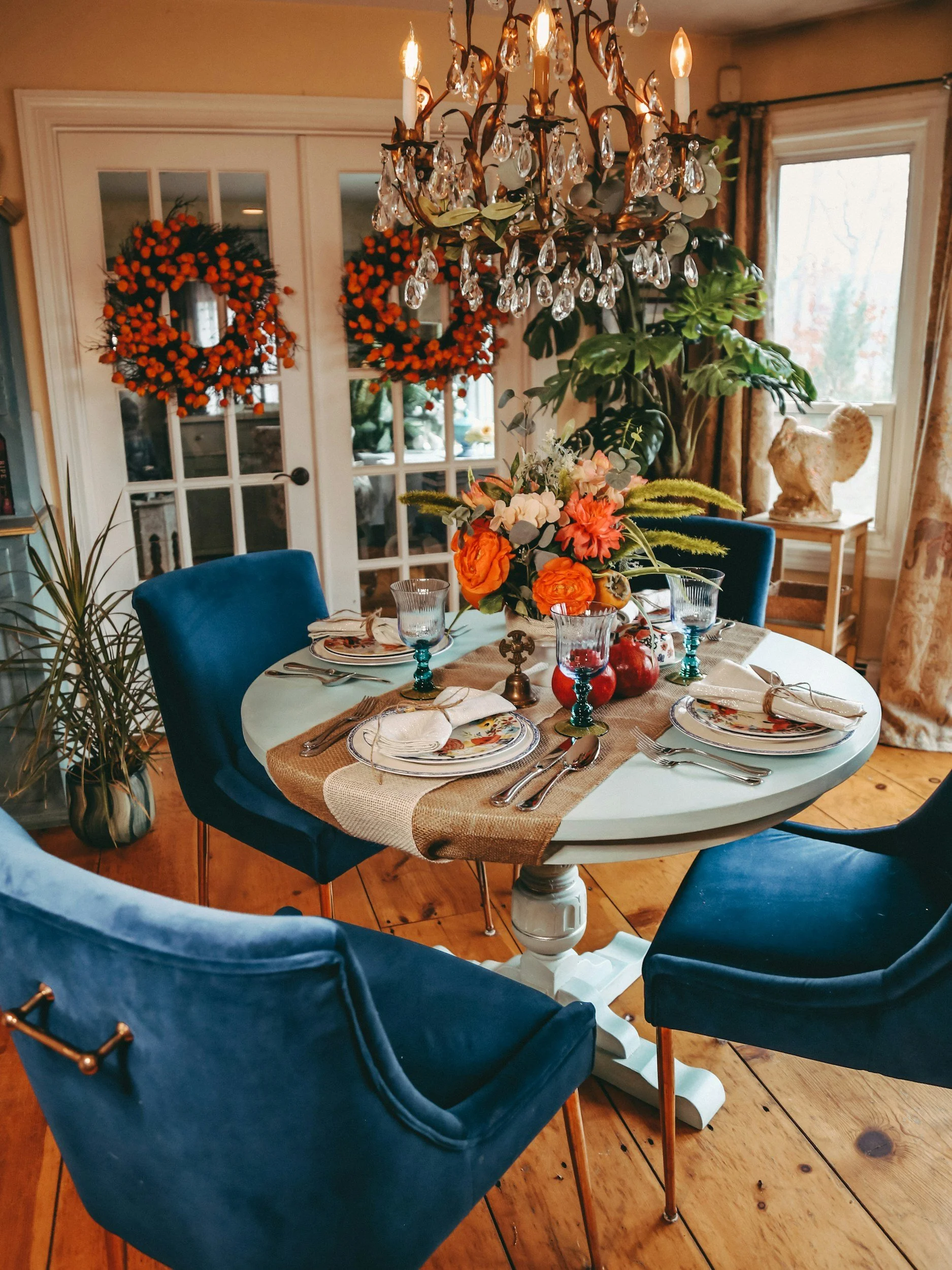 A round dining table set for four, with a floral centerpiece and decorative tableware, surrounded by four blue upholstered chairs, in a cozy room with a chandelier, large windows, and Halloween wreaths on double doors.