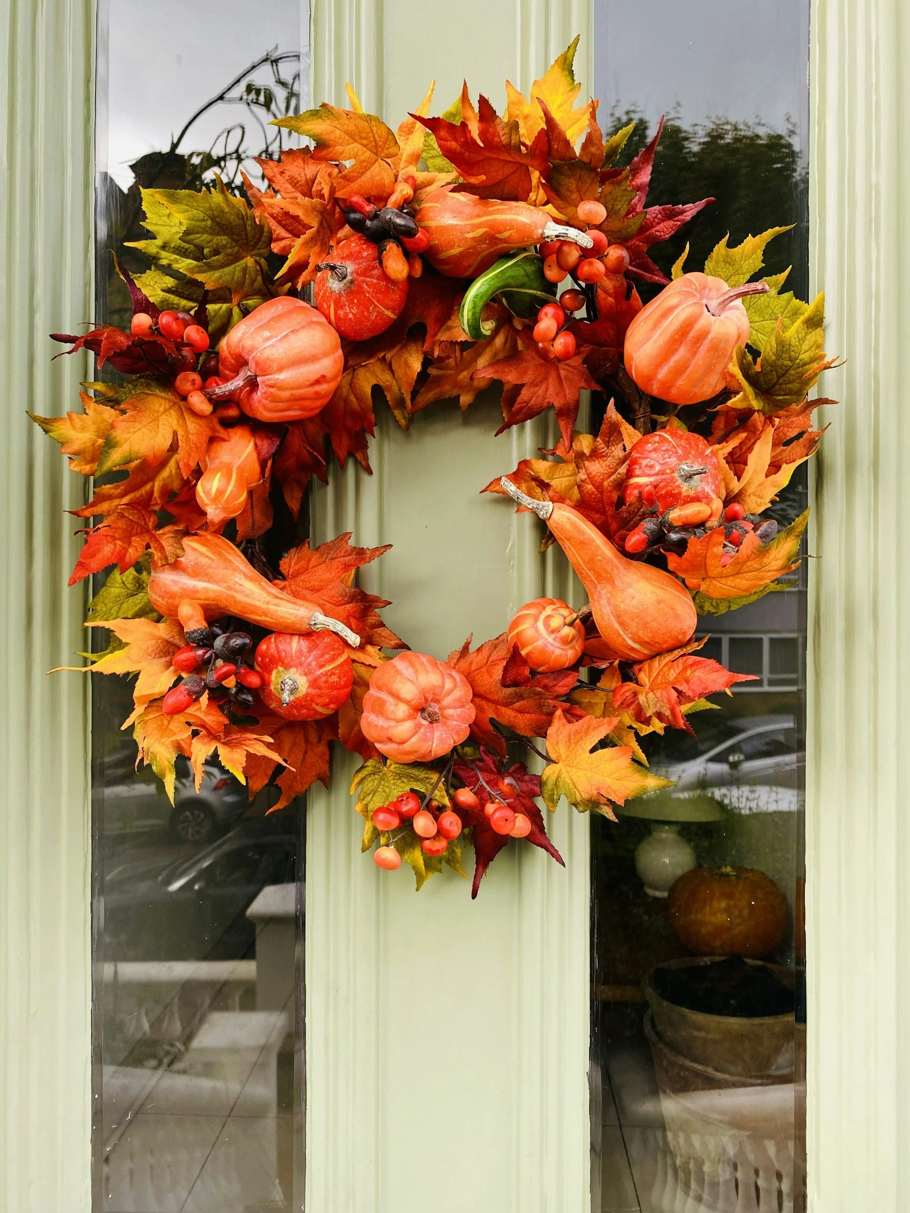 Fall-themed wreath with orange pumpkins, red and orange leaves, berries, and decorative gourds hanging on a green door.