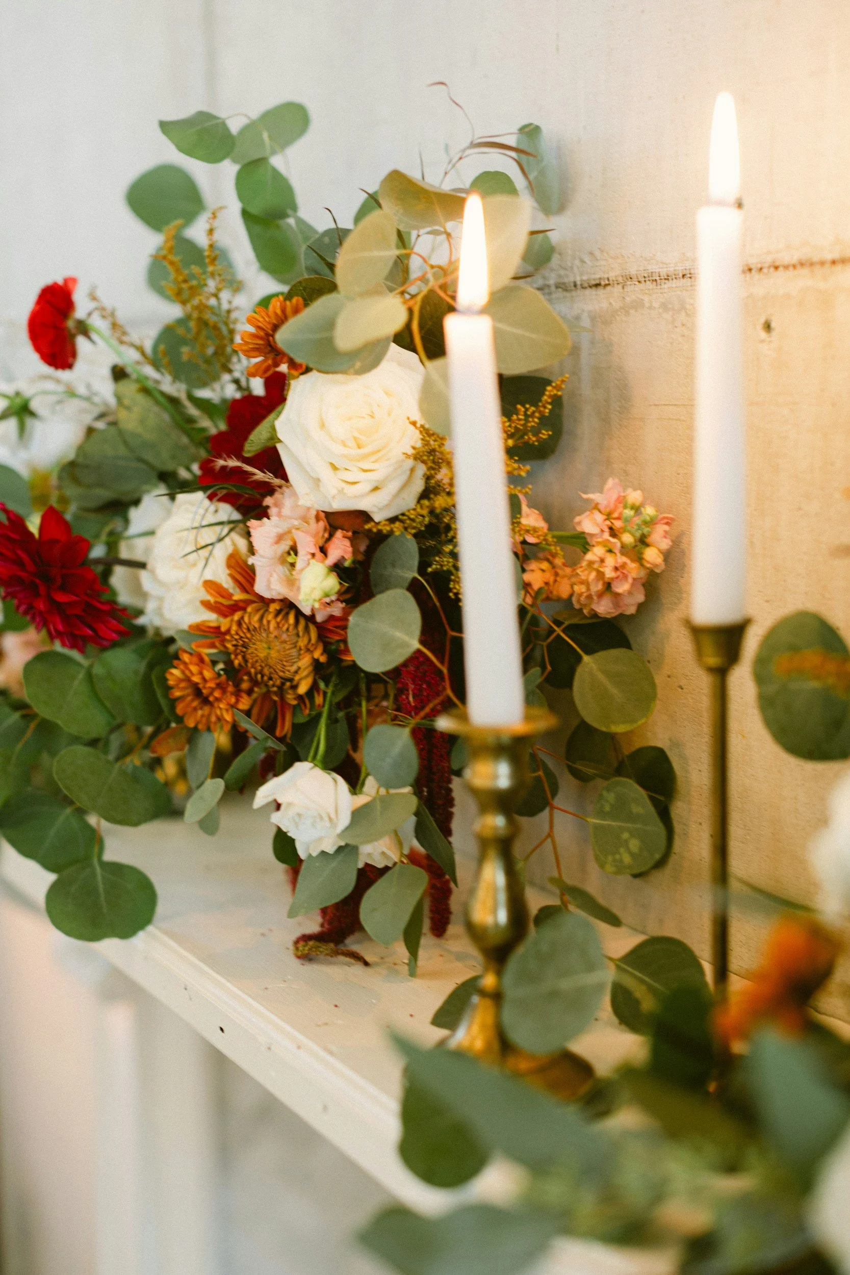A floral arrangement with white roses, red and orange flowers, and green eucalyptus leaves, lit by two white candles in brass holders against a neutral wall.