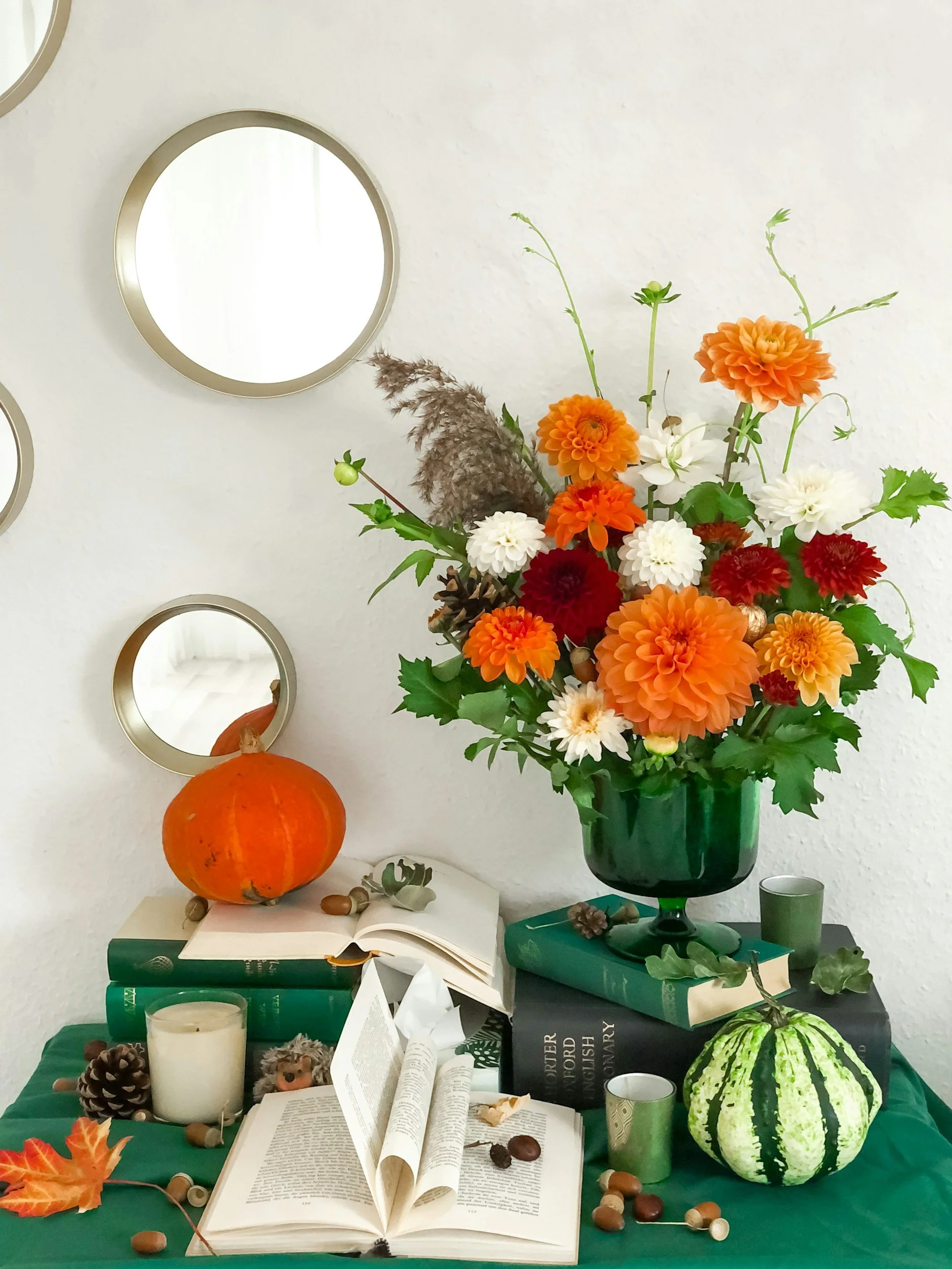 Table decorated with a pumpkin, candles, books, a floral arrangement in a green vase with orange, white, and red flowers, and miscellaneous fall-themed items including pine cones, acorns, and leaves. There are also small cups, a watermelon with green