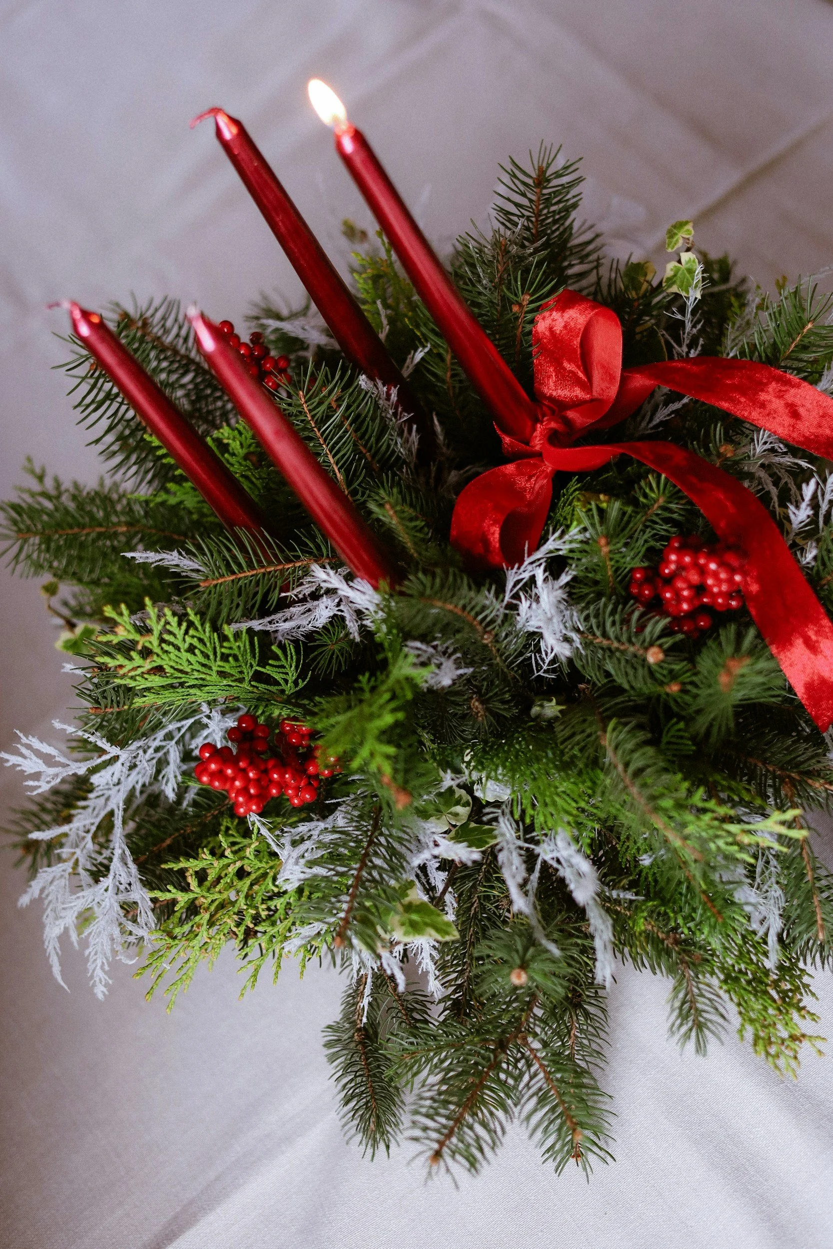 Close-up of an outdoor Christmas wreath with red candles, pine branches, red berries, and a red ribbon, on a gray fabric background.