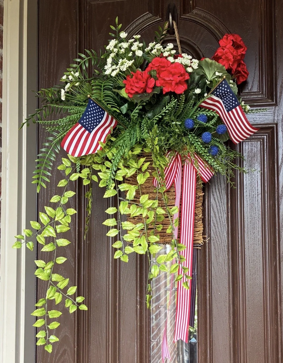 Decorative wreath hanging on a brown wooden door with red hydrangeas, white baby's breath, green foliage, and two small American flags, accented with red, white, and blue ribbons.