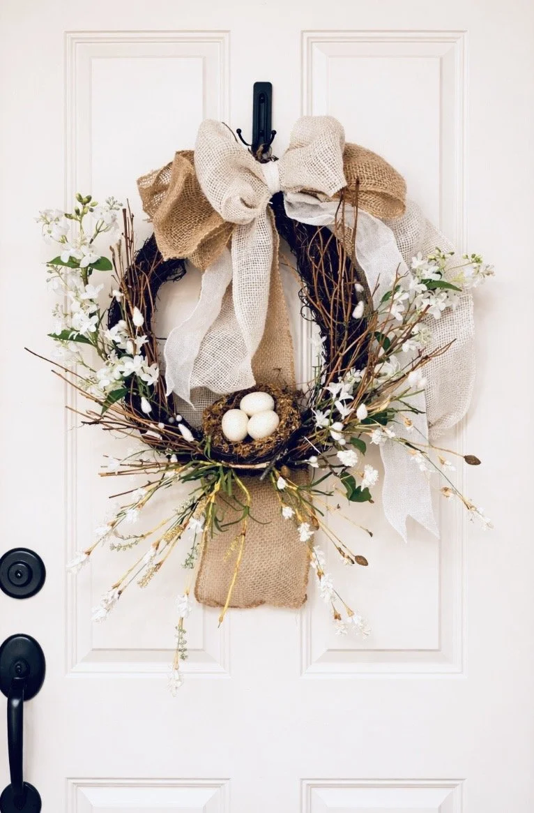 Spring decorated door with a wreath made of twigs, white flowers, and burlap ribbons, featuring three white eggs in a nest at the center.
