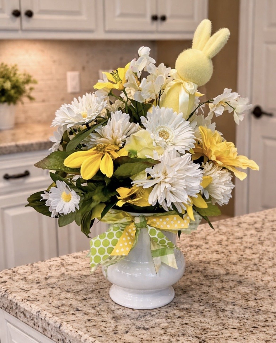 A floral arrangement in a white vase with yellow and white flowers, including daisies and lilies, decorated with a yellow and green polka-dot and checkered bow, placed on a kitchen countertop.