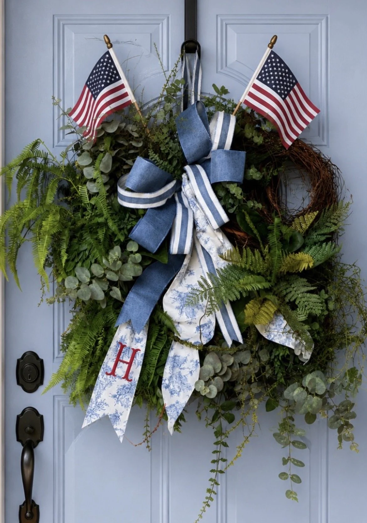 Decorative door wreath with greenery, blue and white ribbons, and two small American flags.
