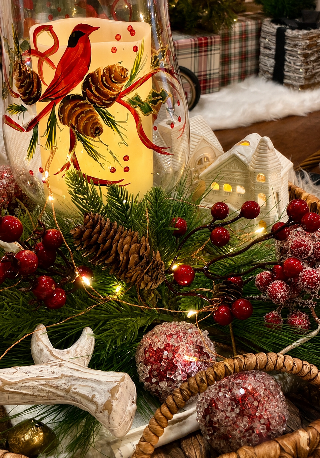 Decorative Christmas centerpiece with a painted candle featuring a cardinal bird, surrounded by pinecones, red berries, evergreen branches, and Christmas ornaments, with festive gift boxes and ceramic houses in the background.