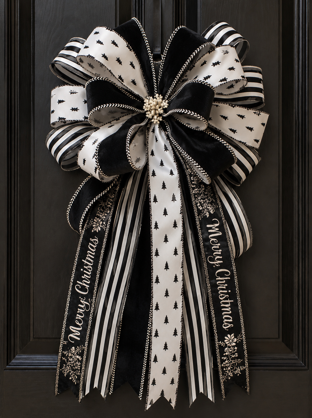 Black and white Christmas wreath with striped, polka dot, and pine tree patterns, featuring a center flower-like cluster of white berries, decorated with ribbons that have 'Merry Christmas' written on them, hanging on a black door.
