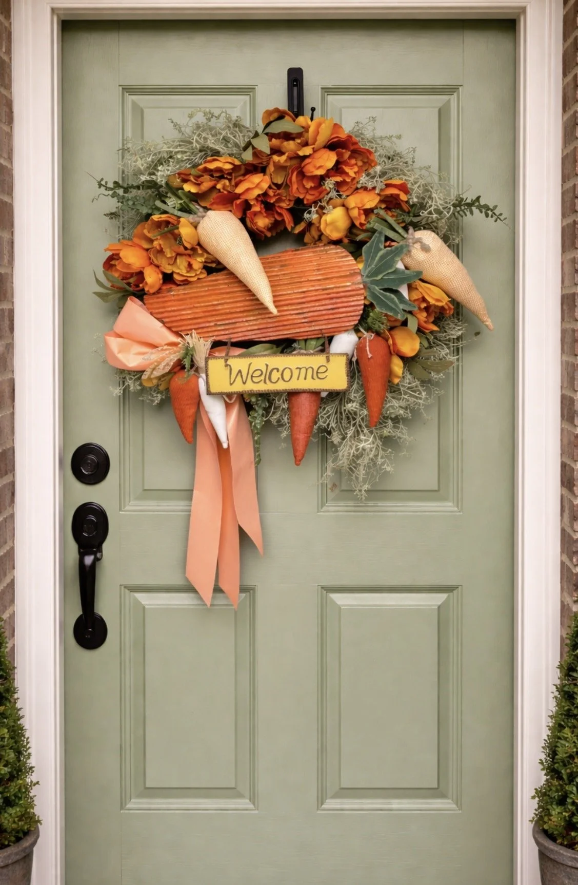 Decorated front door with a spring or autumn-themed wreath made of artificial flowers, carrots, and a yellow sign that says 'Welcome', flanked by two potted plants on either side.