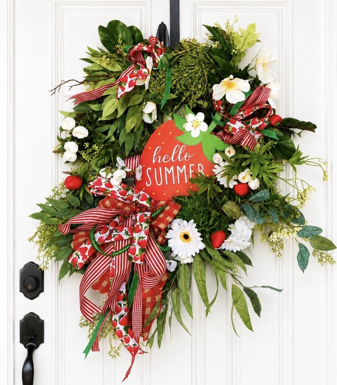 Decorative summer-themed wreath with green foliage, red and white ribbons, white flowers, strawberries, and a red sign that says "hello SUMMER" hanging on a white door.