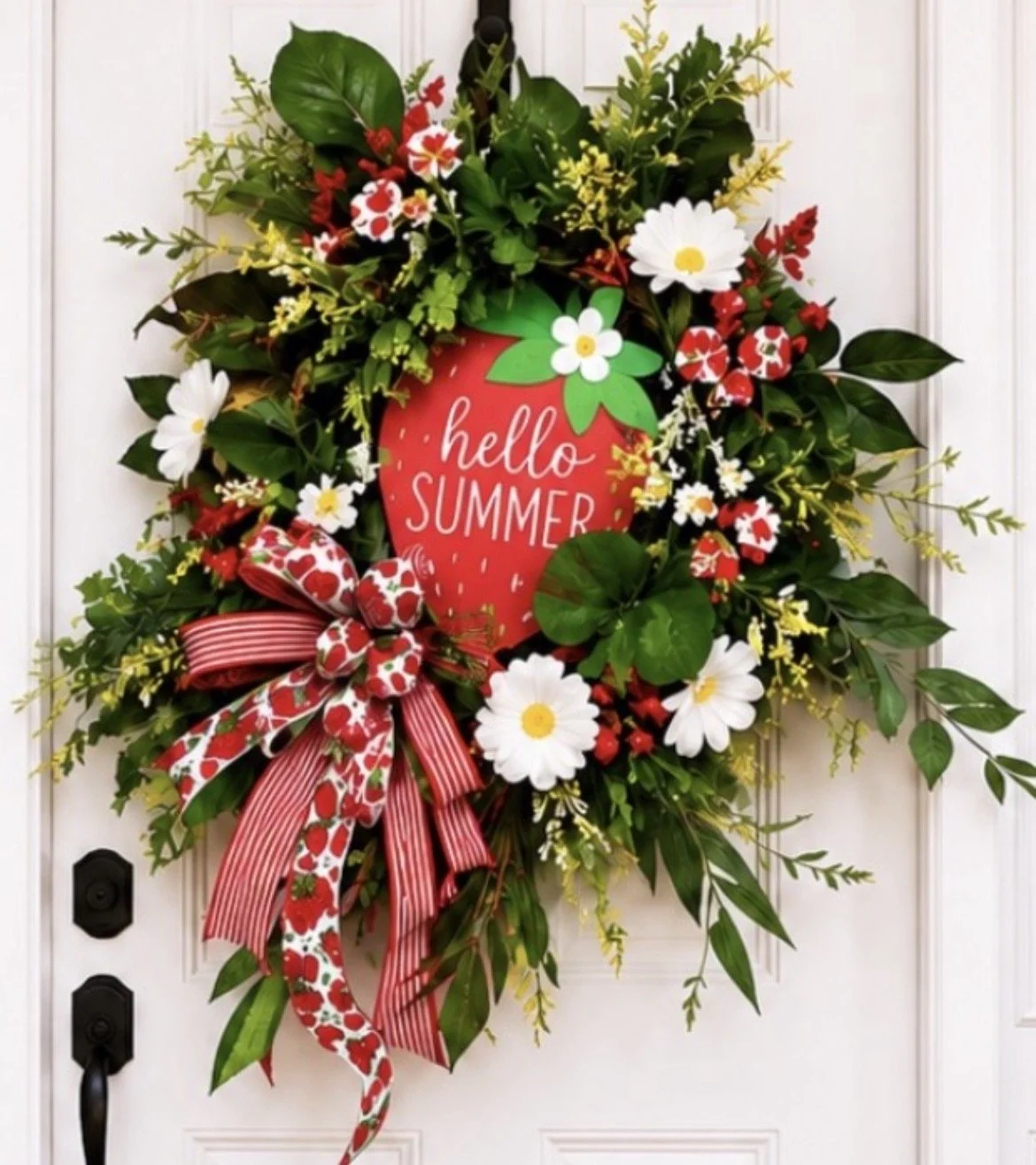 A decorative summer door wreath with green leaves, white daisies, and red and white flowers, featuring a red strawberry-shaped sign that says 'hello SUMMER' and a red and white striped ribbon.