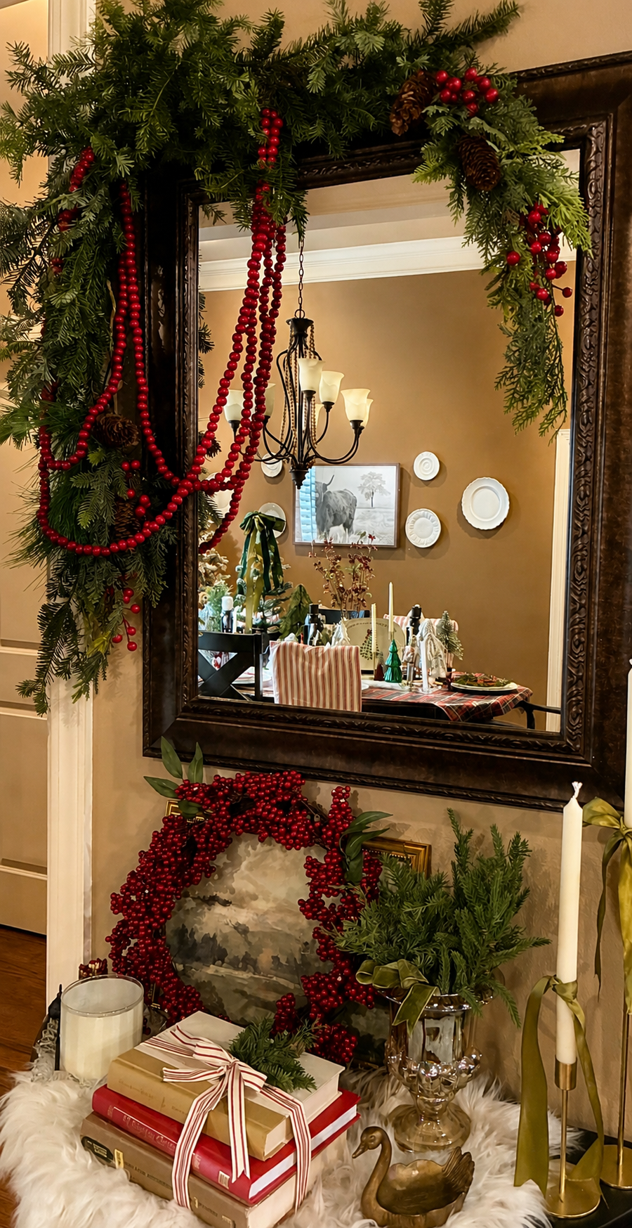 Decorated wall with mirror, holiday greenery, red berry garlands, pinecones, and candles, reflecting a dining room with a painting of an elephant and a festive table setting.