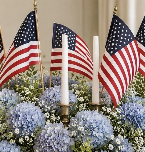 American flags and white candles arranged among blue hydrangeas and small white flowers.