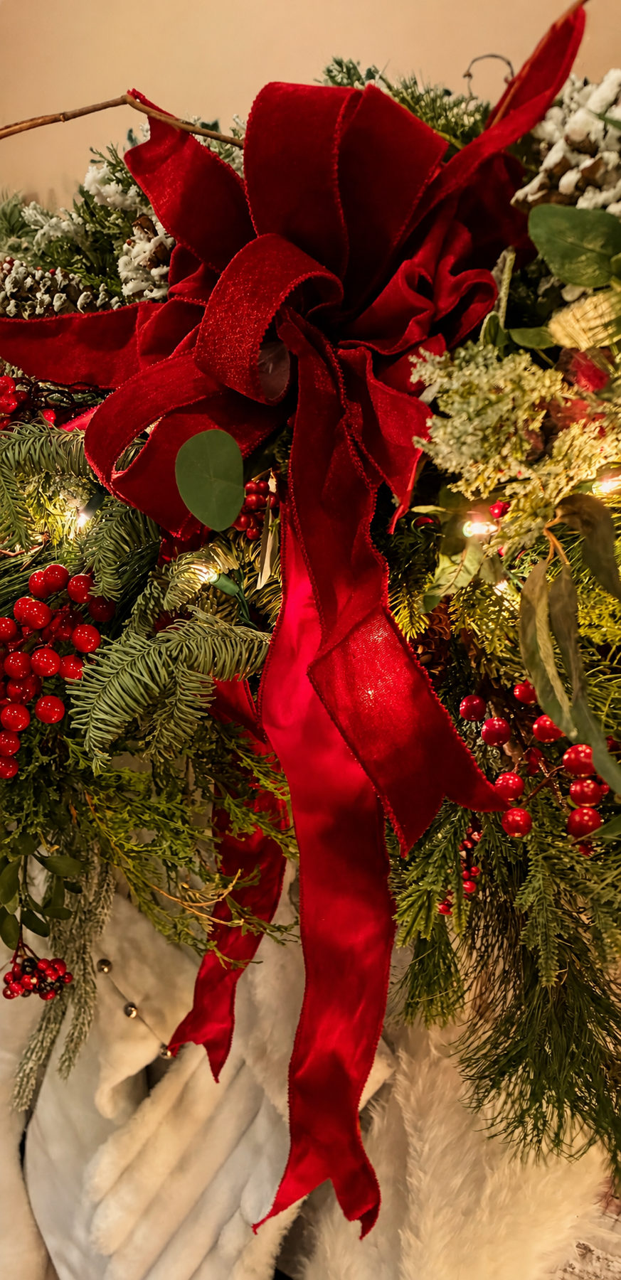 Close-up of a Christmas wreath decorated with a large red velvet bow and red berries, surrounded by green pine branches and white accents.