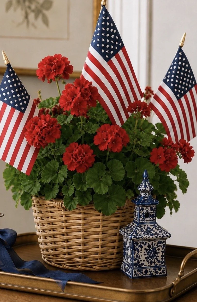 A wicker basket filled with red geraniums and American flags, placed on a wooden tray next to a blue and white decorative lantern.