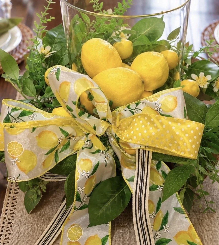A decorative glass vase filled with fresh lemons and lemon branches, adorned with a yellow ribbon with lemon and floral patterns, placed on a table with a linen tablecloth and surrounded by lemon-themed decorations.