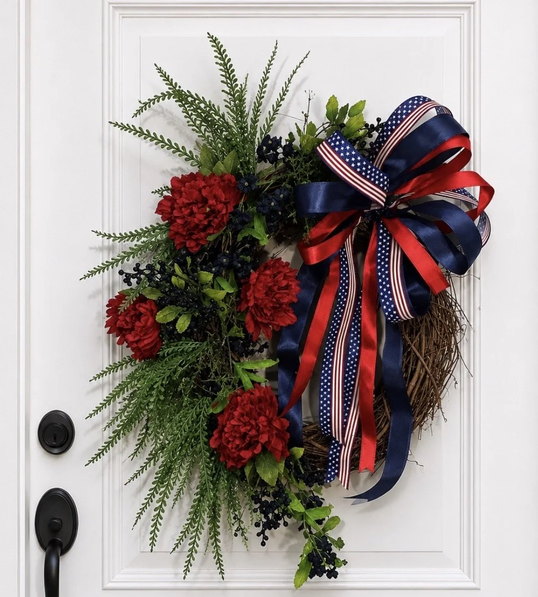 Patriotic door wreath with red flowers, green foliage, and red, white, and blue ribbons.