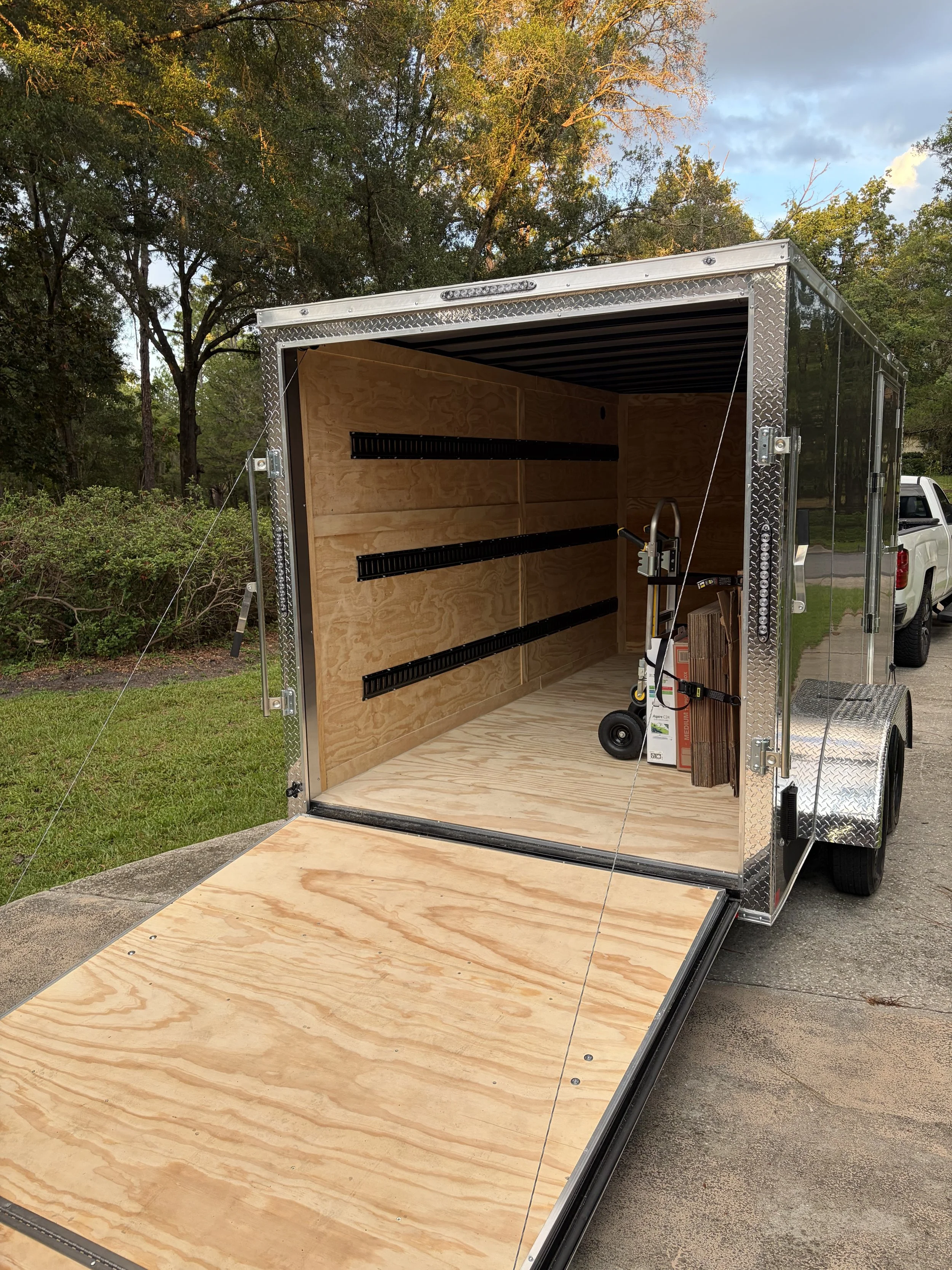 An empty enclosed cargo trailer with a wooden interior, black metal shelving, and a wood ramp door, parked on a driveway with trees and a blue sky in the background.