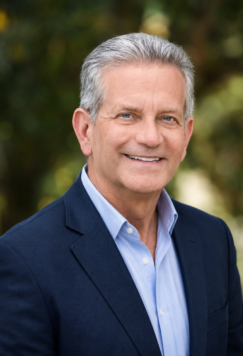 Headshot of Douglas Lundrigan, a middle-aged man with gray hair, blue eyes, wearing a navy blazer and light blue collared shirt, smiling outdoors.